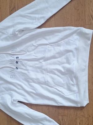 Vit hoodie från Balmain Paris - Säljer en vit hoodie från Balmain Paris med svart text på bröstet och klassisk känguruficka. Huvan har vita dragsnören och insidan är svart. Perfekt för dig som gillar stilrena och exklusiva plagg.