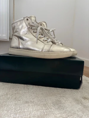 Ralph Lauren sneakers - Snygga high top sneakers i metallic silver med vita snören och gummisula. Skorna har en stilren design med diskreta sömmar och metalliska detaljer som ger en cool vibe. Perfekta för dig som vill sticka ut lite extra i vardagen.