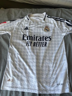 Real Madrid   - Säljer en vit Real Madrid matchtröja från Adidas med subtilt mönster, klubbmärke på bröstet och svarta detaljer. Tröjan har rund hals, korta ärmar och trycket 'Emirates Fly Better' på framsidan. Perfekt för fotbollsträning eller att bära till match.