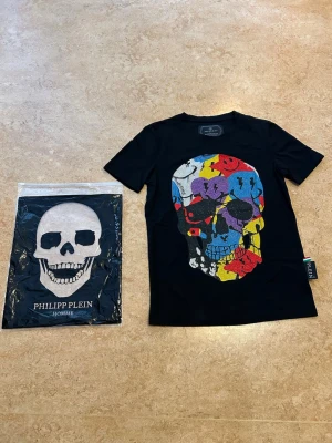 Svart Philipp plein - Säljer en svart t-shirt från Philipp Plein med en stor, färgglad döskalle framtill. Trycket har inslag av blått, rött, gult, vitt, lila och svart. T-shirten har korta ärmar och en klassisk passform. Storlek medium men sitter som en xs. Helt ny aldrig använd