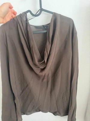 Brun långärmad blus med draperad krage - Snygg brun blus med lång ärm och draperad krage. Tillverkad i ett tunt, lätt och något transparent material som ger en luftig känsla. Perfekt att styla med jeans eller kjol för en trendig look.