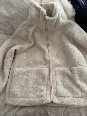 Beige teddyjacka med dragkedja - Mysig beige teddyjacka med hög krage och hel dragkedja framtill. Jackan har två stora fickor framtill och är gjord i fluffigt, mjukt teddy-material som håller dig varm. Perfekt för kyliga dagar och enkel att matcha med olika outfits!!
