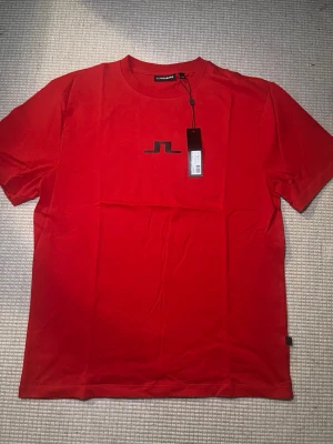 Röd t-shirt med logga från J.Lindeberg - Snygg röd t-shirt från J.Lindeberg med svart logga på bröstet. Klassisk rund halsringning och korta ärmar. Tillverkad i mjuk bomull för skön känsla och stilren look. Perfekt till jeans eller shorts för en clean outfit.