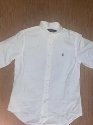 Vit skjorta från Polo Ralph Lauren - Klassisk vit skjorta från Polo Ralph Lauren med blå broderad logga på bröstet. Skjortan har knappar framtill, button-down krage och lång ärm. Perfekt till många olika stilar och enkel att matcha.