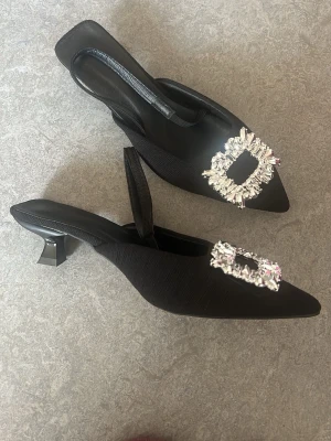 Svarta pumps - Säljer ett par svarta pumps med spetsig tå och öppen häl. Skorna har låg klack. Materialet ser ut att vara textil med en lyxig finish. Perfekta för dig som vill sticka ut lite extra. De är endast använda 1-2 gånger, har en mycket mycket god skick!