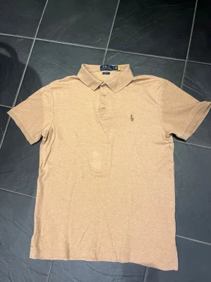 Beige pikétröja från Polo Ralph Lauren - Snygg beige pikétröja från Polo Ralph Lauren i slim fit-modell. Klassisk krage, korta ärmar och broderad logga på bröstet. Perfekt till jeans eller shorts för en clean och stilren look. Obs ngt liten i storleken.