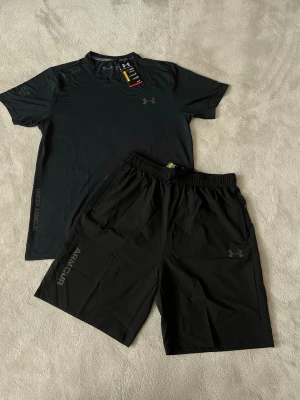 Svart träningsset från Under Armour - Säljer ett svart träningsset från Under Armour med en kortärmad t-shirt och matchande shorts. Båda plaggen har diskreta loggor och är tillverkade i ett lätt och snabbtorkande material som passar perfekt för träning och sportaktiviteter.