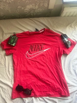 Röd Nike t-shirt med logga - Röd t-shirt från Nike med stor logga och swoosh på bröstet. Klassisk passform och rund halsringning, perfekt för en sportig och avslappnad stil. Tillverkad i mjuk bomull som känns skön mot huden. Pris kan diskuteras.