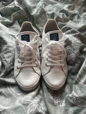 Vita låga sneakers från Jack Marine - Snygga vita sneakers från Jack Marine med klassisk låg profil. Skorna har vita snören, metallhål för snörningen och en vit sula med vågmönster undertill. Ovandelen är i tyg och insidan har randigt foder. Perfekta för en clean och stilren look.
