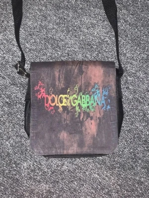 Dolce & Gabbana axelväska med regnbågstryck - Cool axelväska från Dolce & Gabbana i mörkbrunt tyg med regnbågsfärgat tryck på locket. Väskan har justerbar axelrem och fyrkantig form, perfekt för dig som vill sticka ut med en unik accessoar.