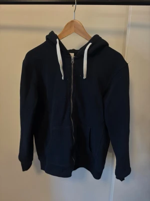 Mörkblå zip hoodie med vita snören - En enkel och stilren mörkblå hoodie med dragkedja framtill och vita snören i huvan. Hoodien har två fickor på magen och ribbade muddar vid ärmslut och nederkant. Perfekt för en avslappnad och bekväm look.