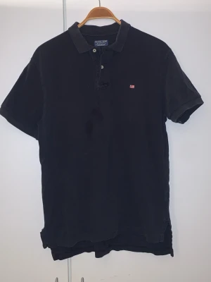 Svart pikétröja från Polo Jeans Company - Klassisk svart pikétröja från Polo Jeans Company by Ralph Lauren. Tröjan har korta ärmar, krage och knappar framtill. Liten röd logga på bröstet och tillverkad i bomull för en bekväm känsla.