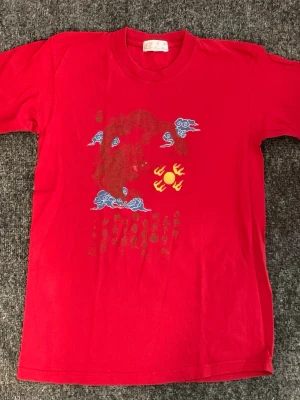 Röd t-shirt med tryck från Fu Min - Röd t-shirt från Fu Min med unikt tryck framtill i blått, gult och rött. Motivet har asiatiska influenser med moln och symboler. Klassisk rund hals och korta ärmar. Perfekt för dig som vill sticka ut med en färgstark look.