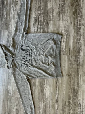 Grå hoodie från Replay med tryck - Snygg grå hoodie från Replay med stort REPLAY-tryck på bröstet och texten 'Denim Goods Since 1981'. Klassisk känguruficka framtill och huva. Perfekt för en avslappnad och trendig look. Mjuk bomullsblandning och långa ärmar.