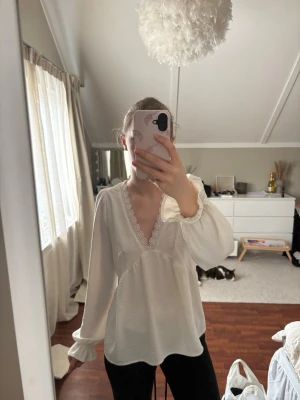 Vit blus med spetsdetaljer - Söt vit blus med djup v-ringning och fina spetsdetaljer längs urringningen. Blusen har långärmade puffärmar med resår vid ärmslut och lätt rynkad midja för en feminin siluett. Tillverkad i ett mjukt, lätt material som ger ett luftigt intryck. Storlek M men skulle säga att den passar S-Xs bättre