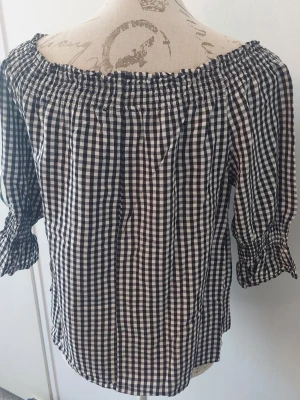 Svartvit offshoulder blus med rutmönster - Trendig svartvit blus med rutigt mönster och offshoulder-modell. Blusen har trekvartsärm med resår och volangdetalj vid ärmslut. Tillverkad i ett lätt och luftigt tyg som ger en avslappnad känsla. 