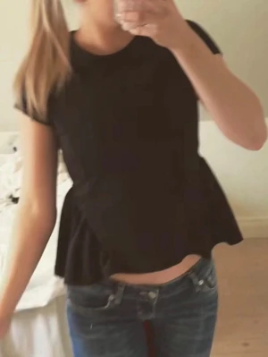 Svart topp från gina - Säljer en svart peplum topp med korta ärmar och rund hals. Toppen har en volang nedtill som ger en snygg och trendig siluett. Perfekt att matcha med jeans eller kjol för en cool look.