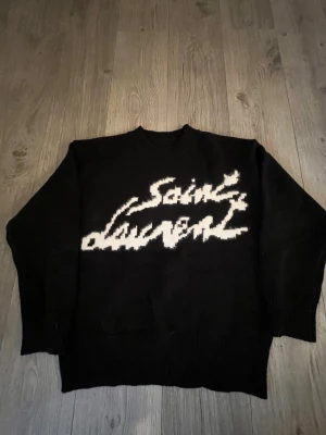 Svart stickad Saint Laurent-tröja - Svart stickad tröja från Saint Laurent med vit, grafisk logotyp framtill. Tröjan har rund halsringning, långa ärmar och ribbstickade muddar. Perfekt för dig som gillar streetstyle och exklusiva märken.