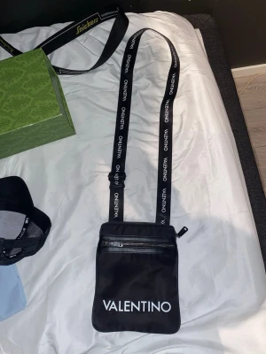 Svart axelväska från Valentino - Snygg svart axelväska från Valentino med justerbart axelband där märkesnamnet står tryckt i vitt. Väskan har en dragkedja upptill och en extra ficka framtill. Tillverkad i ett slätt, tåligt material med minimalistisk design och tydlig logga framtill.