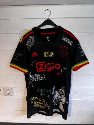 Ajax x Bob Marley limited edition tröja - Unik svart Ajax x Bob Marley matchtröja från Adidas med färgglada detaljer i rött, gult och grönt på ärmarna. Tryck med Ziggo-logga, Ajax-emblem och Bob Marley-motiv samt citat och illustrationer över hela tröjan. Perfekt för fotbollsfans som gillar reggae och streetstyle.