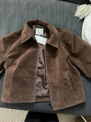 Mocka jacka - Snygg brun mocka jacka med rak passform och bred krage. Jackan har en stilren design med dragkedja framtill och två diskreta sidofickor. Perfekt för dig som gillar en klassisk look med modern twist. Från nakd, storlek 36. Aldrig använd! Nypris 1000