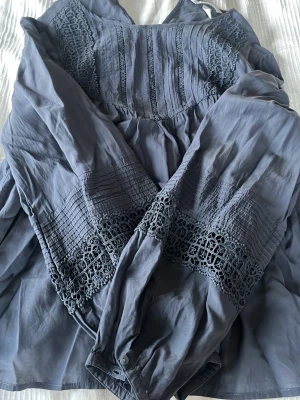 Mörkblå blus med spetsdetaljer - Snygg mörkblå blus från H&M med långa ärmar och vackra spetsdetaljer på både ärmar och över bröstet. Blusen har en lös passform och knappar vid ärmsluten. Tillverkad i ett tunt, mjukt tyg som känns skönt mot huden. Storlek 42/L