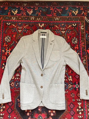 Pier One kavaj - Stilren ljusgrå kavaj från Pier One med två knappar framtill, två stora framfickor och en bröstficka. Kragen är i en klassisk notch modell med ett litet ankare. Materialet är mjukt och bekvämt, därav en mer avslappnad look. Knappt använd. Nypris ligger runt 500 kr.