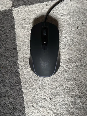 Svart gamingmus med tribal-mönster - En svart gamingmus med sladd och ett stilrent tribal-liknande mönster på ovansidan. Musen har två sidoknappar och ett scrollhjul. Ergonomisk form och matt yta för bra grepp, perfekt för gaming eller datorarbete.