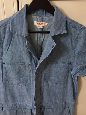 Ljusblå denim jumpsuit Soaked - Snygg ljusblå denim jumpsuit från Soaked med korta ärmar, två bröstfickor och knappar framtill. Jumpsuiten har krage och dragsko i midjan för en avslappnad men cool look. Perfekt till dig som gillar en trendig och bekväm stil. Använts endast få gånger.