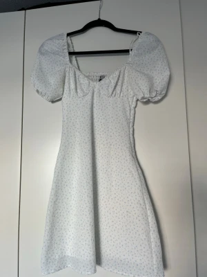 Vit blommig kortklänning med puffärm - Söt vit kortklänning med små blå blommor och puffärmar. Klänningen har en smockad rygg, fyrkantig urringning och lätt utställd kjol. Perfekt för sommarens alla tillfällen och ger en romantisk vibe.