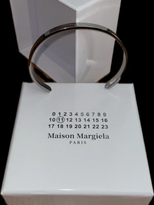 Maison Margiela silverfärgat armband - Snyggt och stilrent armband från Maison Margiela i silverfärgad metall. Armbandet är stelt och har märkets logotyp ingraverad på ovansidan. Perfekt accessoar för dig som gillar minimalistisk och modern design.