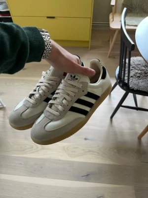 Adidas Samba OG vita och svarta sneakers - Klassiska Adidas Samba OG sneakers i vitt och svart med beige mockadetaljer och gummisula. Skorna har de ikoniska tre svarta ränderna på sidan, vit ovandel i läder och beige snörning. Perfekta för en clean och retro stil. Helt nya, använda utomhus 1 gång.
