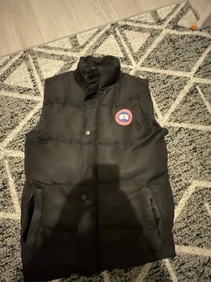 Svart dunväst från Canada Goose - Snygg svart dunväst från Canada Goose med hög krage och två fickor framtill. Västen har det klassiska runda Canada Goose-märket på bröstet och stängs med knappar. Perfekt för lager-på-lager och håller dig varm under kyliga dagar.
