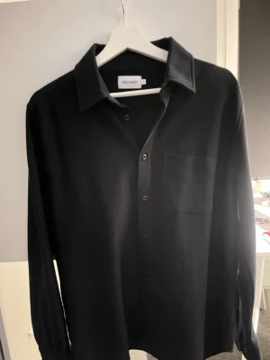 Svart skjorta från East West - Klassisk svart skjorta från East West med lång ärm och bröstficka. Skjortan har knappar framtill och en stilren, enkel design som passar till det mesta. Perfekt för dig som gillar en clean och tidlös look.