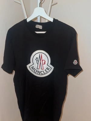 Svart Moncler t-shirt med logga - Svart t-shirt från Moncler med stor broderad logga på bröstet och mindre logga på ärmen. Klassisk rund halsringning och korta ärmar. Tillverkad i mjuk bomull som känns skön mot huden. Perfekt för en stilren och trendig look.