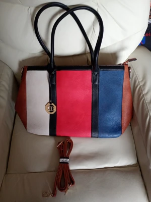 Flerfärgad handväska med breda ränder - Snygg handväska från Dudlin i konstläder med breda vertikala ränder i vitt, rött och blått på framsidan samt bruna sidor och baksida. Väskan har svarta handtag, guldfärgad logodetalj och avtagbar axelrem. Perfekt för dig som gillar färg och stilren design.