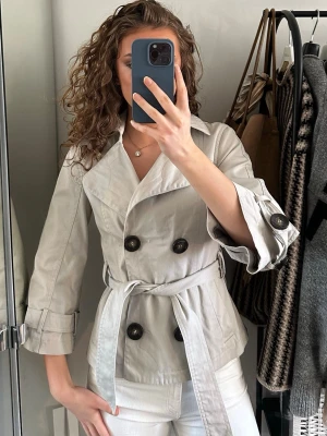 Beige kort trenchcoat med knytskärp - Stilren beige trenchcoat med dubbelknäppning och stora svarta knappar. Jackan har ett matchande knytskärp i midjan och uppvikta ärmslut. Klassisk krage och lätt figursydd passform. Perfekt för vår och höst, tillverkad i ett tunt bomullsliknande material.