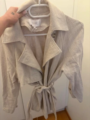 Beige trenchcoat från bubbleroom - Stilren beige trenchcoat från bubbleroom i ett lätt, strukturerat tyg. Jackan har klassisk krage, knytband i midjan och långa ärmar. Perfekt för dig som gillar minimalistisk och tidlös stil.