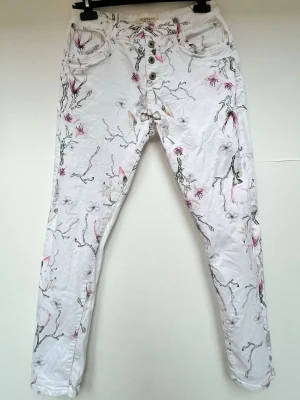 Vita jeans - Vita jeans med blommigt mönster i storlek L/40 ifrån märket Jewelly - 98% bomull, 2% elastan - Har några skavanker, vilket syns på de två sista bilderna: längst ner på vänster bakben är det en mycket svag fläck. Uppe på vänster baksida är det även en söm lös. - En silver stjärn på höger bakficka