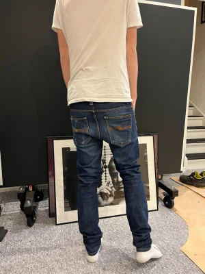 Blå slim fit jeans från Nudie Jeans - Säljer ett par mörkblå slim fit jeans från Nudie Jeans med klassiska fem fickor och slitningar på bakfickorna. Jeansen har en snygg tvätt med ljusare partier framtill och kontrastsömmar. Perfekta för en avslappnad och trendig stil.