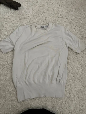 Vit kortärmad topp från ZARA - En enkel och stilren vit topp från ZARA med korta ärmar och rund hals. Toppen har ribbade detaljer vid ärmslut och nederkant, vilket ger en snygg passform. Perfekt att matcha med jeans eller kjol för en clean look. Använd cirka 2 gånger 