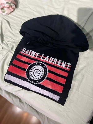 Svart Saint Laurent hoodie - Svart hoodie från Saint Laurent med stor huva och ett fett tryck framtill i rött, vitt och svart. Texten 'SAINT LAURENT' och ett SL-emblem med stjärnor och lagerkrans ger en sportig vibe. Perfekt för dig som vill sticka ut med en ikonisk streetwear-look. Är orginalt i storlek L men passar som en m 