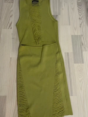 Olivgrön ärmlös klänning med volang - Säljer en olivgrön ärmlös klänning med markerade volangdetaljer framtill och längs sidorna. Klänningen har en kroppsnära passform och en cut-out detalj i midjan. Perfekt för dig som vill sticka ut med en unik look.