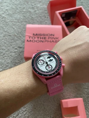 Omega x Swatch Pink Moonphase klocka - Säljer en Omega x Swatch Mission to the Pink Moonphase klocka i rosa med vit urtavla och svarta detaljer. Klockan har biokeramisk boett, rosa armband med Omega-logga och cool månfas-funktion. Perfekt statement piece för dig som gillar färg och unika accessoarer.