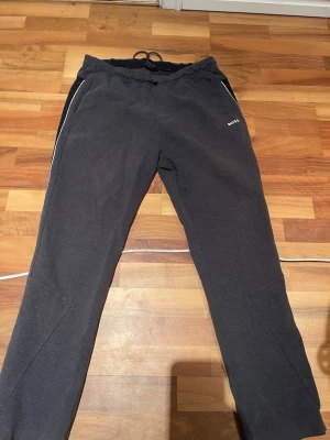 Mörkgrå mjukisbyxor från BOSS - Snygga mörkgrå mjukisbyxor från BOSS med vita detaljer längs fickorna och logga på vänster sida. Byxorna har dragsko i midjan och raka ben. Perfekta för chill dagar eller när du vill ha en avslappnad stil.