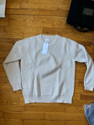 Beige stickad tröja från Maison Margiela - Säljer en stilren beige stickad tröja från Maison Margiela. Tröjan har rund halsringning, ribbade muddar och är gjord i ett mjukt, strukturerat material. Perfekt för dig som gillar minimalistisk och clean stil.