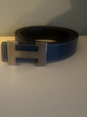 Blått skärp med H-spänne i skinn - Snyggt blått bälte i skinn med kontrastsömmar och ett stort H-format metallspänne. Baksidan är svart och bältet har en stilren design som passar till många outfits. Perfekt accessoar för att lyfta din look.