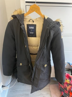 Svart parkasjacka från Svea - Svart parkasjacka från Svea med beige foder och fuskpäls på huvan. Jackan har dragkedja, knappar, två stora fickor framtill och broderad Svea-logga. Perfekt för kalla dagar och riktigt snygg streetstil.