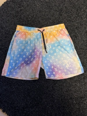 Färgglada Louis Vuitton badshorts - Säljer ett par färgglada Louis Vuitton shorts med pastellfärgat mönster i blått, rosa, gult och vitt. Shortsen har LV-loggor över hela tyget, elastisk midja och svart snörning framtill. Perfekta för sommaren och riktigt snygga till streetwear-stilen.