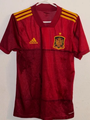 Spaniens matchtröja Adidas röd - Röd matchtröja från Adidas med Spaniens landslagsmärke och en gul stjärna ovanför. Tröjan har gula detaljer på axlarna och Adidas-logga på bröstet. Tillverkad i lätt och ventilerande material, perfekt för fotboll eller träning. Inga hål eller så, den är riktigt bra skick och är direkt från Adidas 
